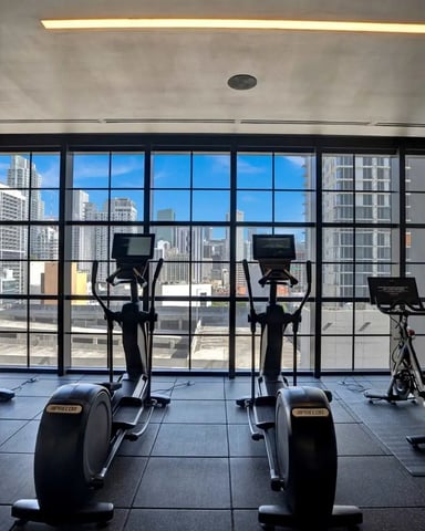 Modern gym with cardio equipment facing floor-to-ceiling windows overlooking city skyline and high-rise buildings