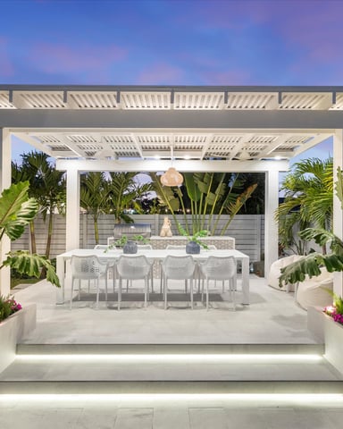 Modern pergola-covered patio with white dining table, tropical plants, and illuminated planters at dusk
