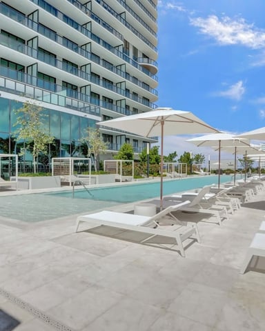 Modern high-rise building with rooftop pool, lounge chairs, umbrellas, and palm trees under blue sky
