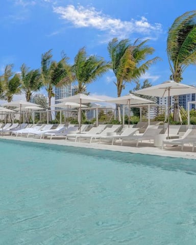 Resort pool with turquoise water, lounge chairs, white umbrellas, palm trees, and modern buildings under blue sky