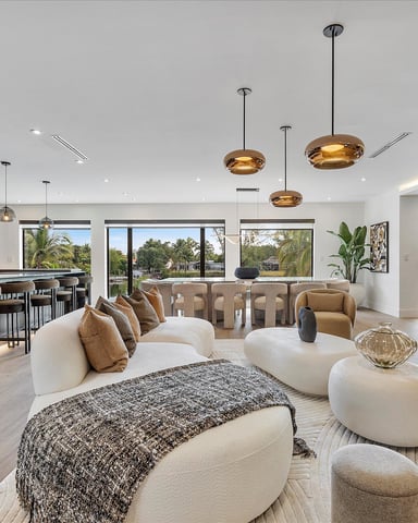 Open-plan living room with cream seating, gold pendant lights, kitchen with dark cabinetry, and views of palm trees through large windows