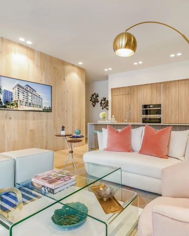 Modern studio apartment with light wood walls, white bed with coral pillows, glass coffee table, arc floor lamp, and open kitchen with wood cabinetry