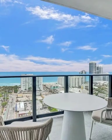 High-rise balcony with round white table, two woven chairs, and panoramic ocean and coastal city views