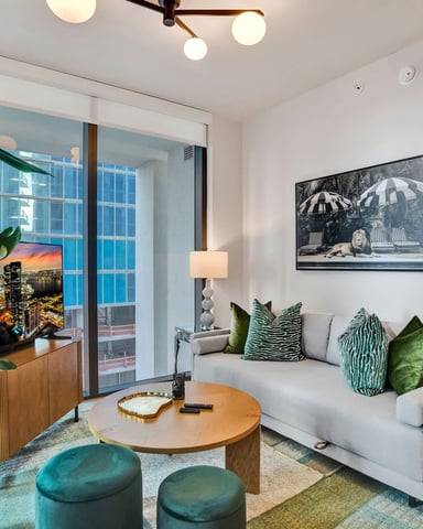 Modern living room with light gray sofa, green patterned pillows, round wood coffee table, teal ottomans, potted plant, and floor-to-ceiling windows