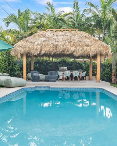 Backyard pool with thatched-roof cabana, lounge chairs, dining area, and palm trees